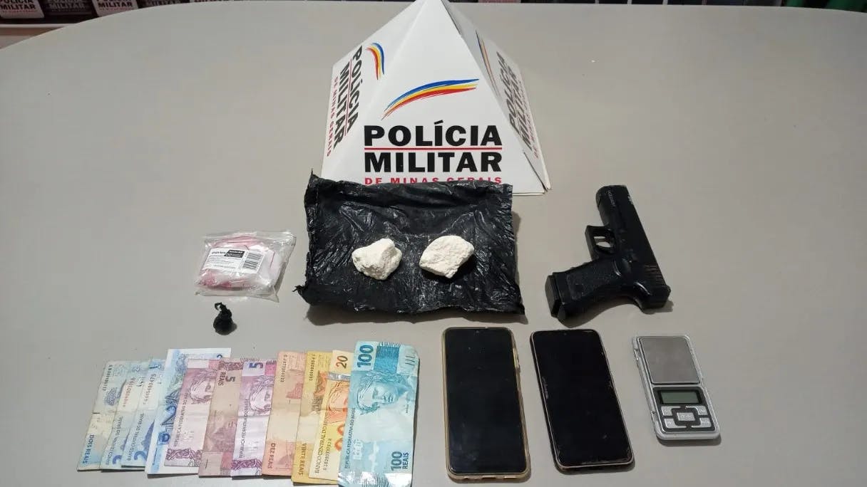 imagem colorida mostrando prisma da Polícia Militar ao lado das duas pedras de cocaína, arma falsa, notas de dinheiro de dois, cinco, dez, e 100 reais, além celulares
