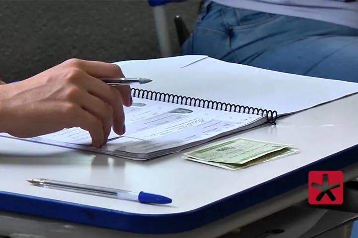 fotografia mostrando uma mão segurando caneta com os dedos encostados no caderno de votação. Ao lado uma outra caneta bic azul e titulo eleitoral em cima de uma identidade. Ambos sobre uma mesa.