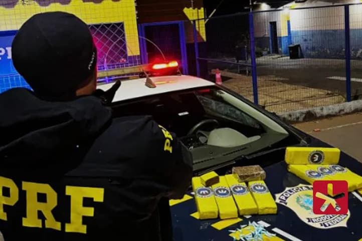 imagem colorida mostrando policial da PRF de costas e a sua frente as barras de cocaína, pinos de maconha e porções de crack sobre o capô de uma viatura da PRF