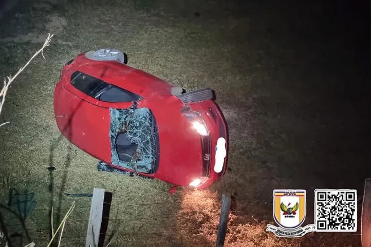 imagem colorida mostrando veículo de cor vermelha, tombado de lado após queda em ribanceira. O carro está bastante danificado em meio a vegetação