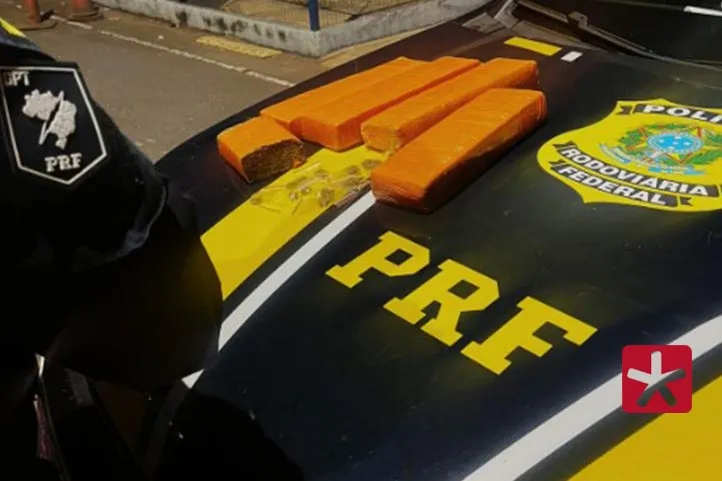 imagem colorida mostrando tabletes de maconha quatro tabletes e meio de maconha, ao lado de vários papelotes de cocaína em cima do capô de viatura da PRF