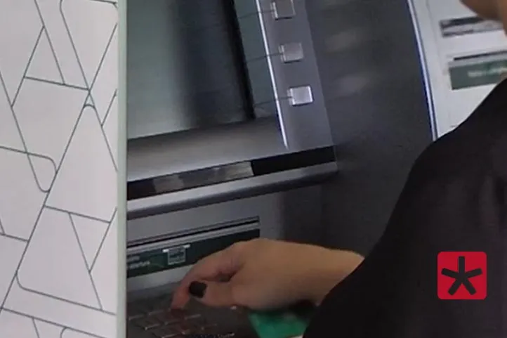 imagem colorida mostrando pessoa de pé em frente a um terminal eletronico bancário acessando conta, com a mão direita sobre o teclado do equipamento.